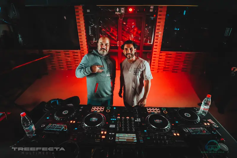 Dos personas posan detrás de la cabina de DJ, iluminadas en rojo